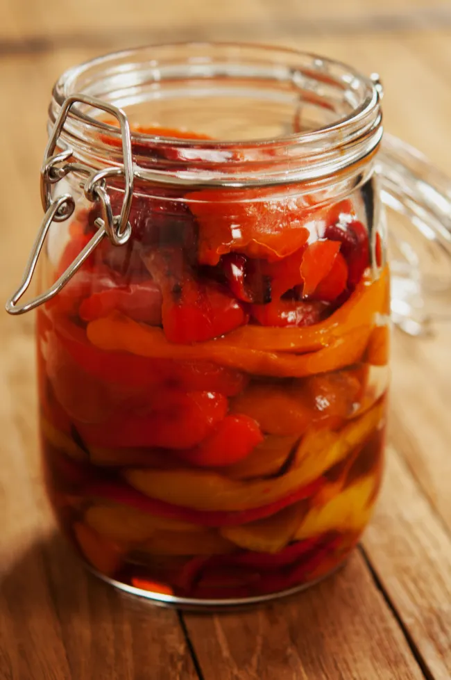 Glasgefäß mit Drahtbügel gefüllt mit Schichten von rot, orange und gelb geröstetem Paprika-Streifen auf einem Holzbrett