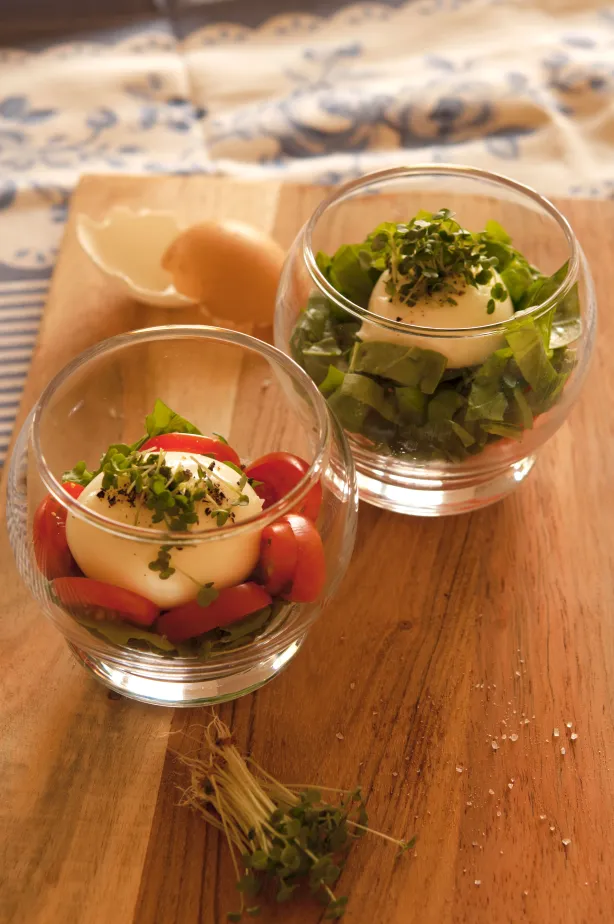Zwei kleine Gläser mit Schichten aus grünem Gemüse, Tomaten und gekochten Eiern auf einem Holzbrett