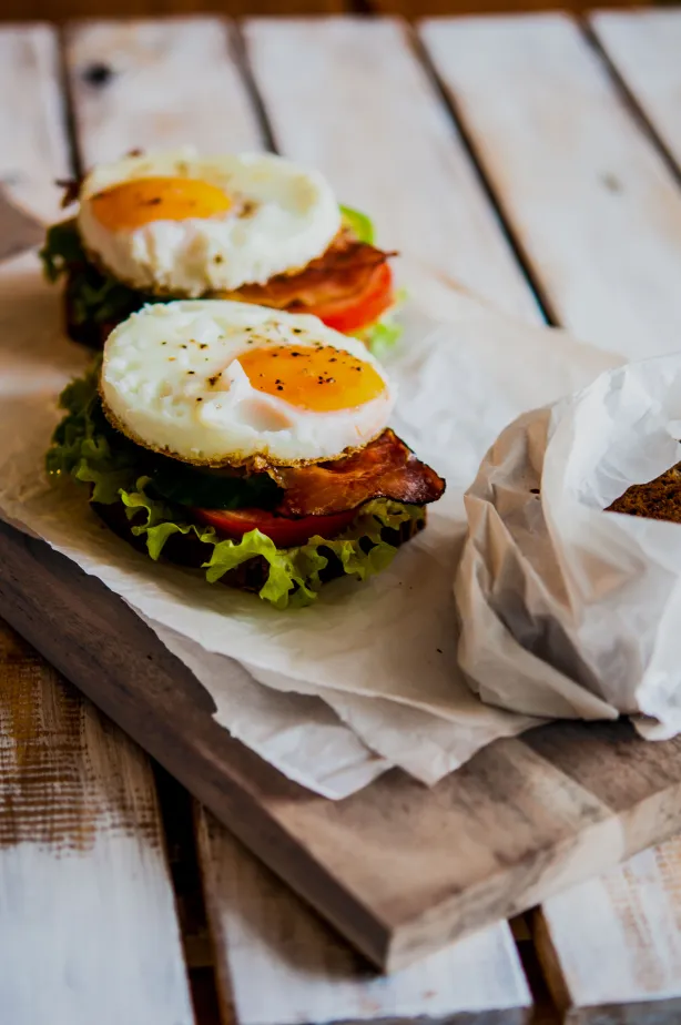 Zwei offene Sandwiches mit Spiegeleiern, Speck, Salat und Tomate auf einem Holzbrett, herzhafte rustikale Stimmung in warmen Brauntönen