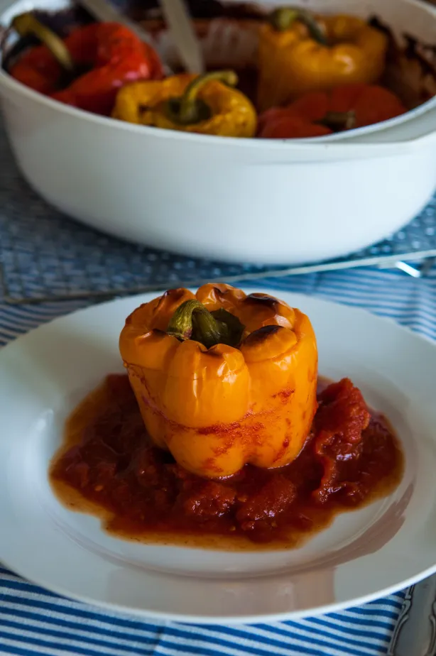 Gefüllte orangefarbene Paprikaschote auf weißem Teller mit Tomatensauce, weitere gefüllte Paprika im Topf im Hintergrund
